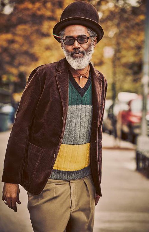 Bearded adult man in glasses, brown bowler hat and corduroy jacket, and colorful sweater walks on city sidewalk in autumn.
