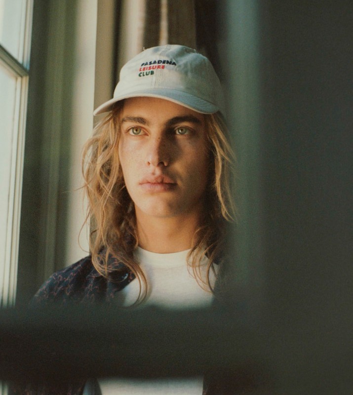 Portrait of a young adult man with long blond hair wearing a white baseball cap, posing indoors near a window with a diffused light.