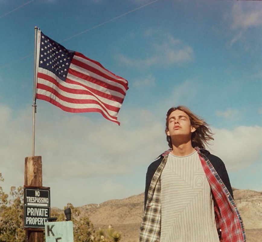 Young man stands against a desert landscape, with an American flag waving in the background against a partly cloudy sky.