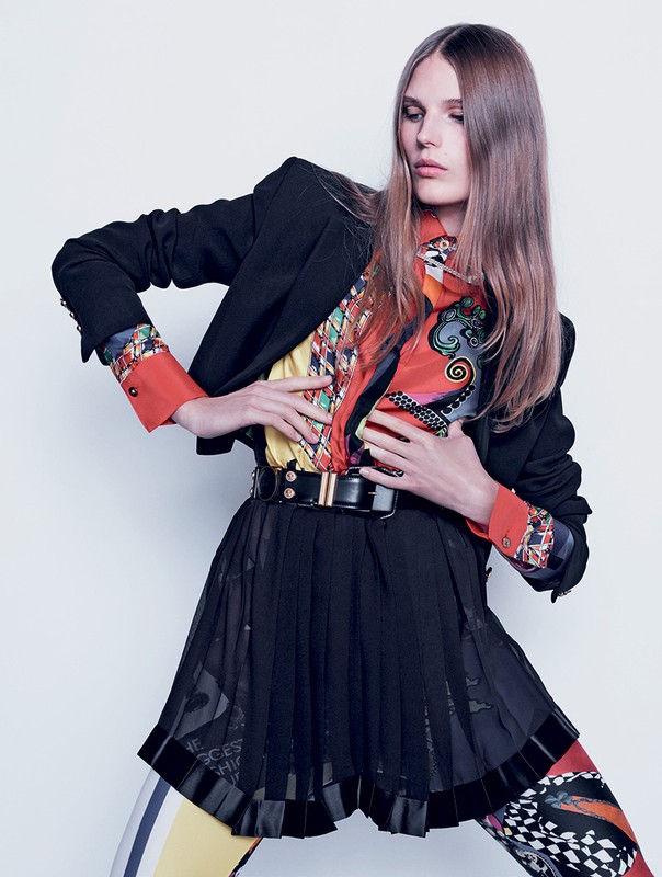 Fashionable woman in a black blazer, colorful patterned top, and black skirt poses against a white background in a studio shot.
