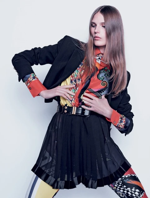Fashionable woman in a black blazer, colorful patterned top, and black skirt poses against a white background in a studio shot.