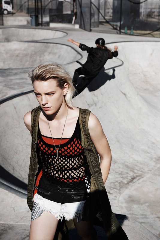 A young adult woman in streetwear poses in the foreground of a skatepark, with another person skating in the background, on a sunny day.