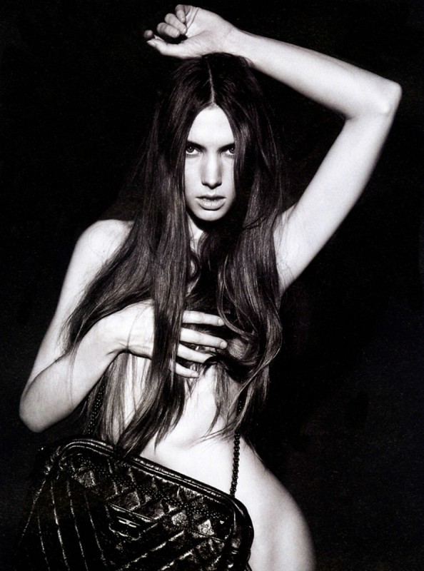 Black and white studio portrait of a topless young woman with long dark hair, a black leather bag, against a dark background.