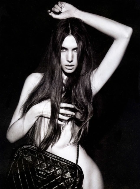 Black and white studio portrait of a topless young woman with long dark hair, a black leather bag, against a dark background.