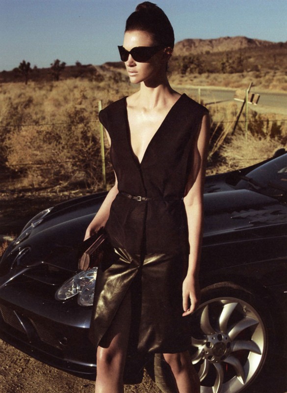 A stylish adult female model in a black dress and sunglasses poses near a sleek black car, with a desert landscape backdrop and a retro aesthetic.