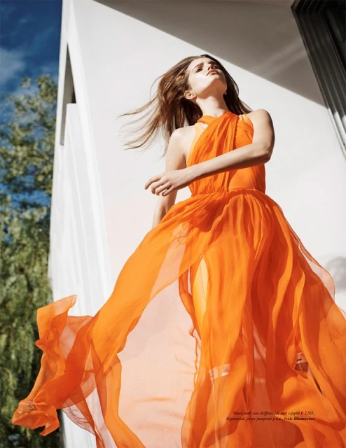 A young woman poses in a flowing orange dress with a modern architectural backdrop, bathed in natural light against a clear blue sky.