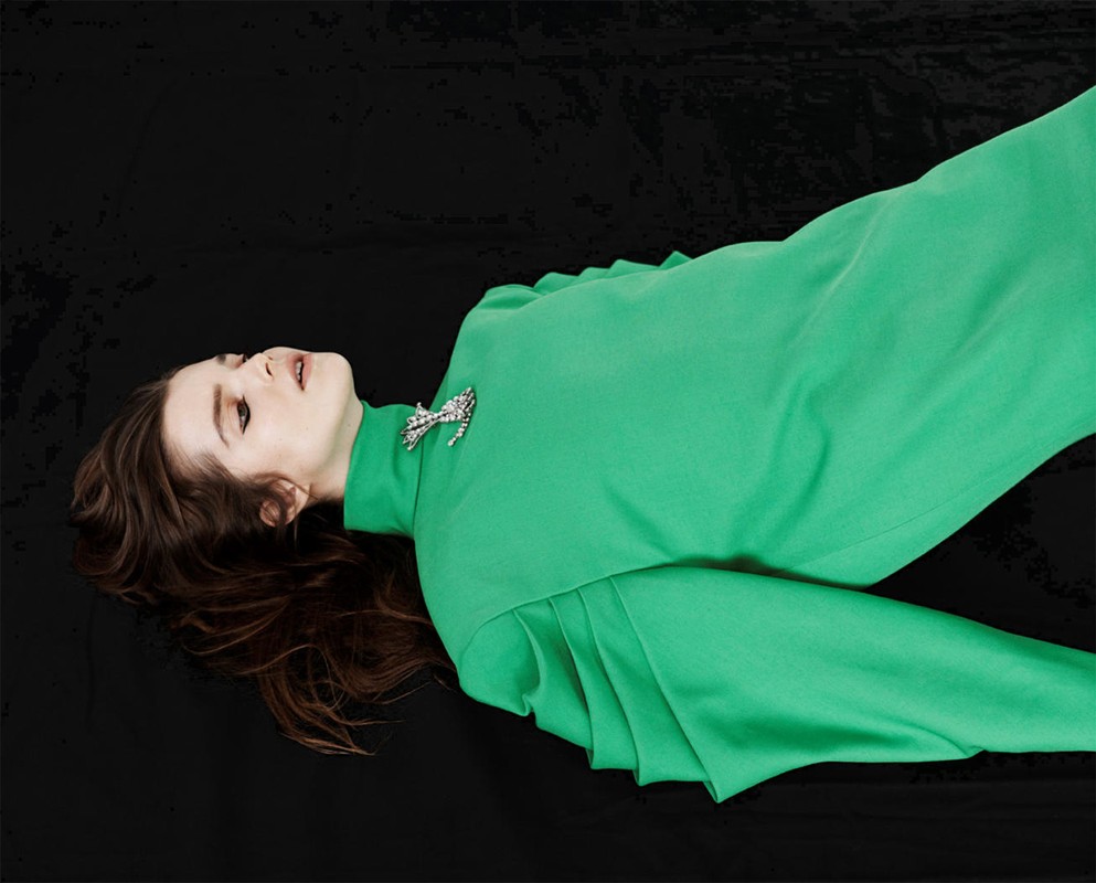 Woman with brown hair poses on a black surface wearing a green dress with a brooch in a studio setting with directional lighting.