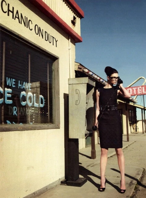 Woman in little black dress using payphone next to mechanic shop with motel in background under blue sky in editorial fashion shot.