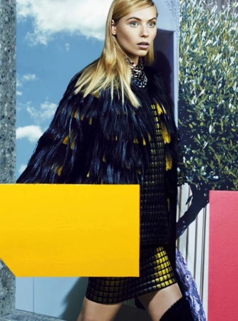 Model with long blonde hair wearing a textured dark cape and patterned outfit against a surreal backdrop with clouds and trees.