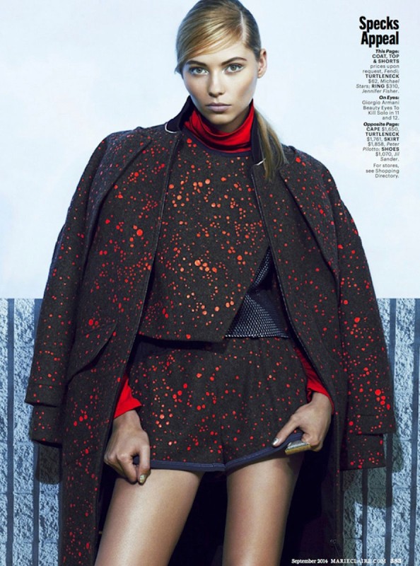 Fashion editorial shot of a woman in a patterned co-ord set and coat with a red turtleneck, posing in front of a textured wall.