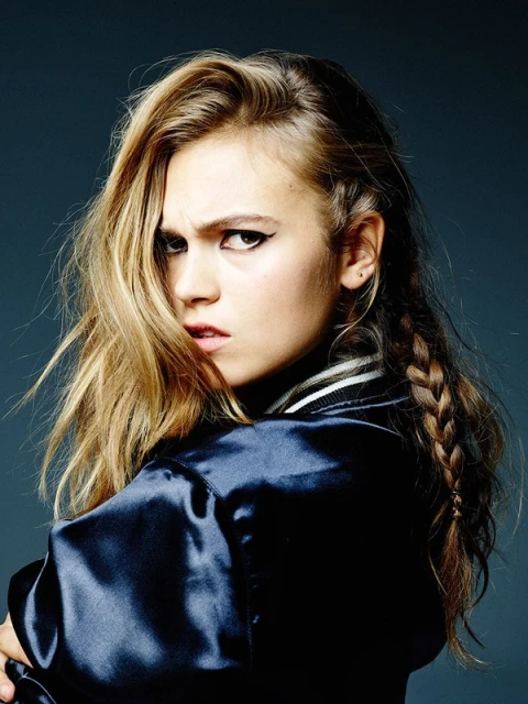 Portrait of a young adult woman with long brown hair and wing eyeliner, wearing a blue satin jacket, looking at the camera.