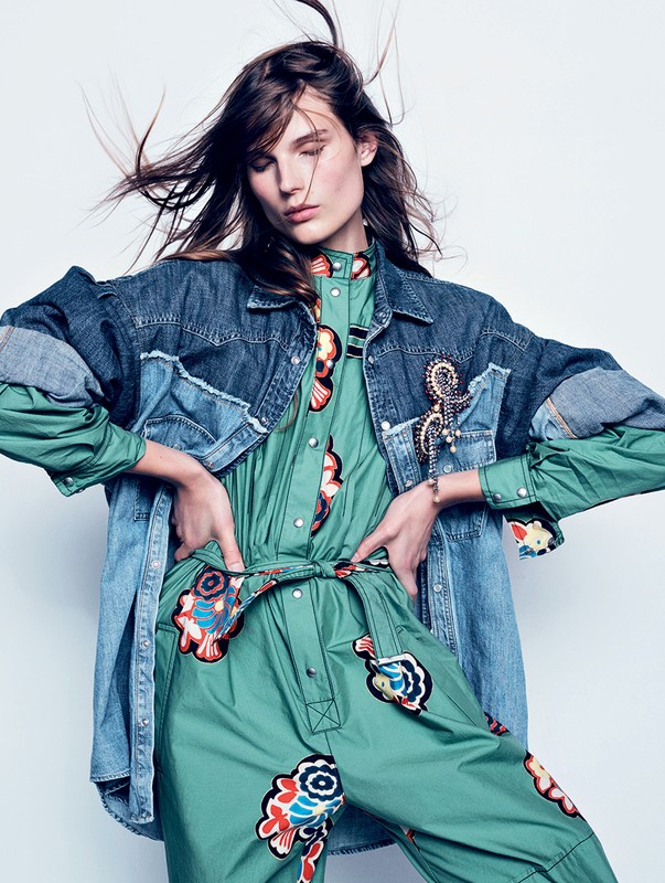 Model with flowing hair wearing a patterned green jumpsuit and oversized denim jacket against white seamless.