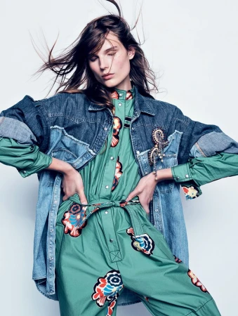 Model with flowing hair wearing a patterned green jumpsuit and oversized denim jacket against white seamless.