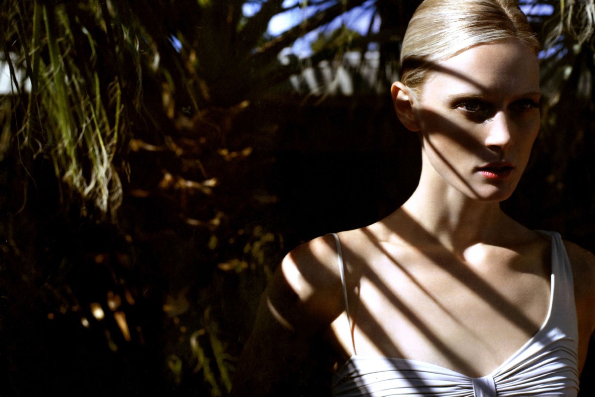 Portrait of a blonde woman wearing a white top with strong shadow patterns cast across her face and body by palm fronds.