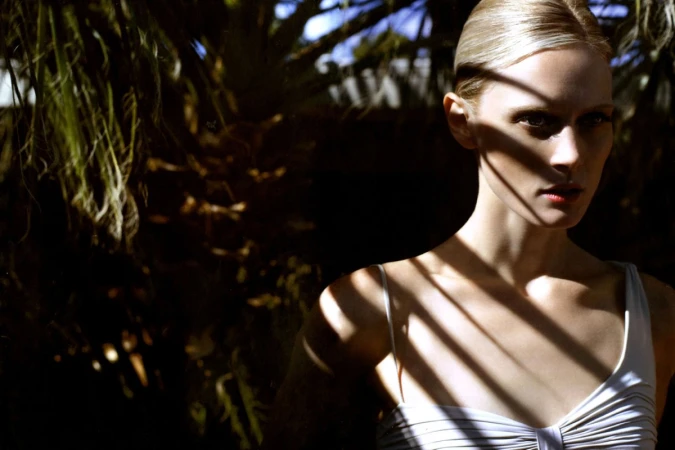 Portrait of a blonde woman wearing a white top with strong shadow patterns cast across her face and body by palm fronds.