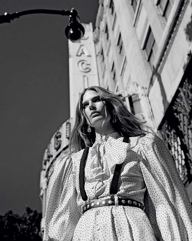 Black and white portrait of an adult female model in a polka-dot dress and suspenders, standing in an urban setting under a street lamp.