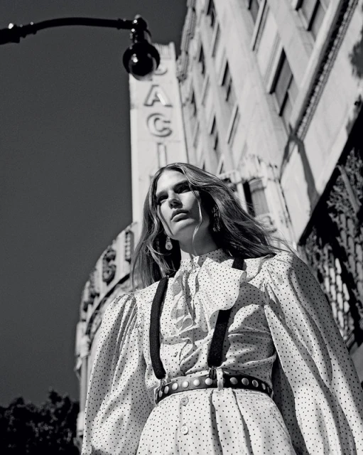 Black and white portrait of an adult female model in a polka-dot dress and suspenders, standing in an urban setting under a street lamp.