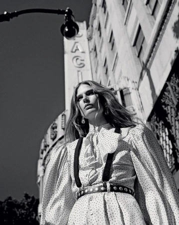 Black and white portrait of an adult female model in a polka-dot dress and suspenders, standing in an urban setting under a street lamp.