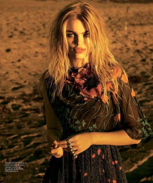A fair-skinned young adult female model with blond hair wears a floral dress while posing on a beach in a fashion editorial photo.