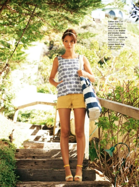 Woman in 60s-inspired fashion posing on wooden steps outdoors. She is wearing a patterned top, yellow shorts, and yellow heeled sandals.