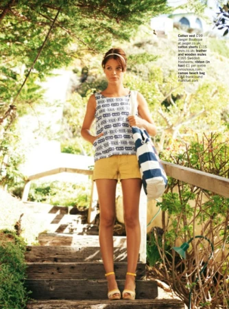 Woman in 60s-inspired fashion posing on wooden steps outdoors. She is wearing a patterned top, yellow shorts, and yellow heeled sandals.