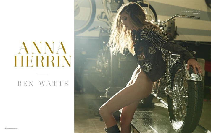 Woman in studded leather jacket poses near a motorcycle in a garage setting with studio lighting, creating a dramatic editorial image.