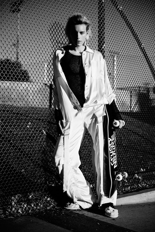 Black and white portrait of a woman in a baggy white jumpsuit and black top, holding a skateboard, leaning against a chain-link fence.