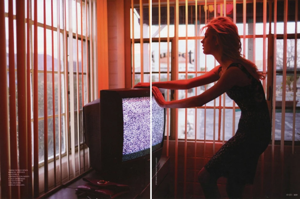 Indoor fashion shot of a woman in a black dress leaning against a television set with window blinds and an orange-toned color grade.