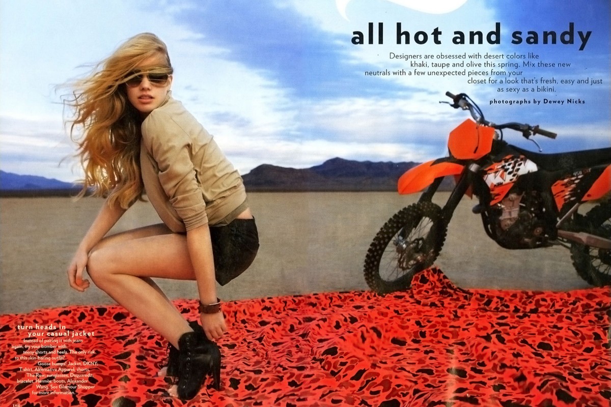 Fashion shot of a woman crouching on patterned fabric with a dirt bike in a desert landscape under a blue sky with copy.