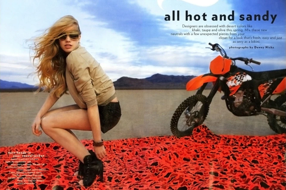 Fashion shot of a woman crouching on patterned fabric with a dirt bike in a desert landscape under a blue sky with copy.