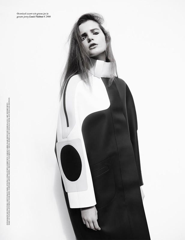 Editorial fashion shot featuring a stylish adult female in a black and white geometric-patterned coat against a bright, neutral backdrop.