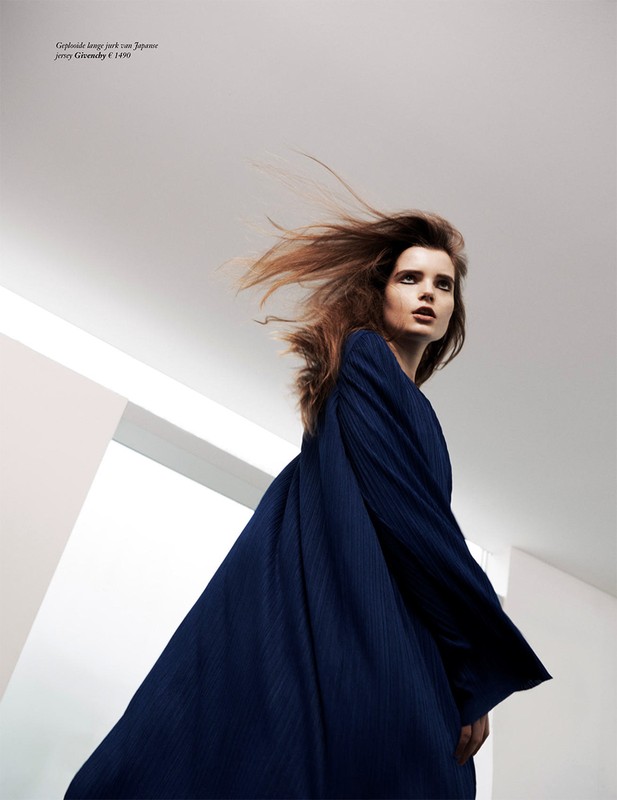 Fashion shot of a female model in a flowing navy dress with long sleeves, standing in a white room with an upward angle.