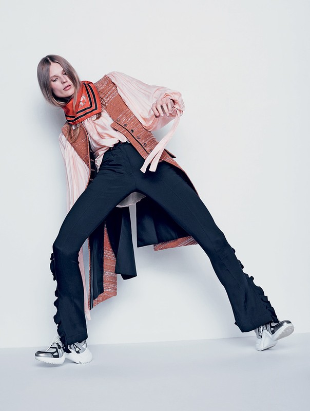 Fashion model wearing a pink blouse, rust vest, black pants with ruffles, and silver sneakers in a full-length studio shot.