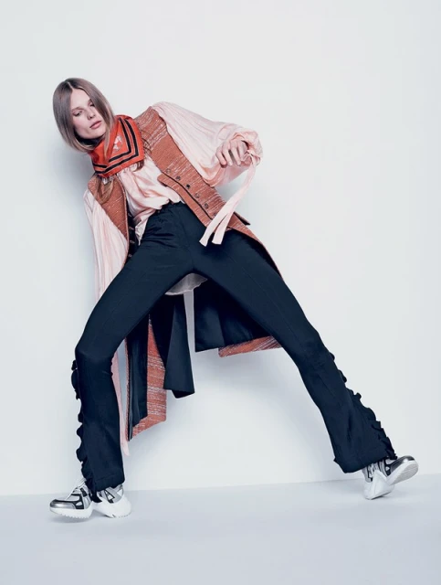 Fashion model wearing a pink blouse, rust vest, black pants with ruffles, and silver sneakers in a full-length studio shot.