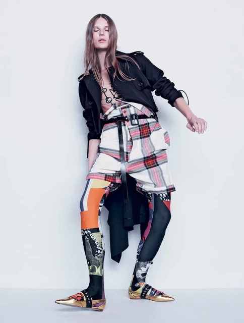 A fashion model poses in a studio wearing an eclectic outfit including a jacket, belted plaid skirt, leggings with graphic prints, and gold shoes.