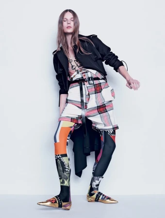 A fashion model poses in a studio wearing an eclectic outfit including a jacket, belted plaid skirt, leggings with graphic prints, and gold shoes.