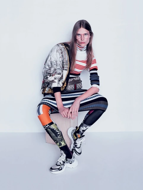 Editorial fashion shot of a woman with glasses, wearing a colorful outfit, striped sweater, printed jacket, and patterned leggings, sitting in a studio.