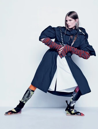 Model in a dark sculptural coat with striped arm warmers and patterned knee socks posing against a white seamless backdrop.