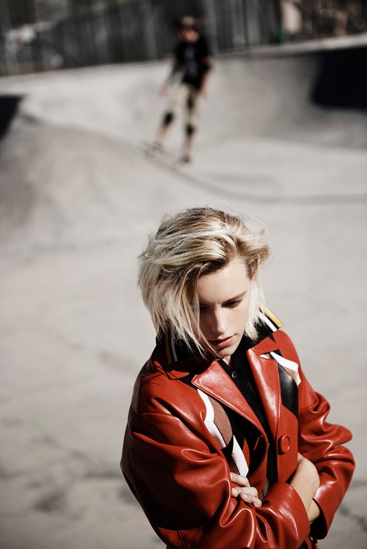Stylish young woman in red leather coat posing at the edge of a concrete skatepark bowl, with skateboarder in background.