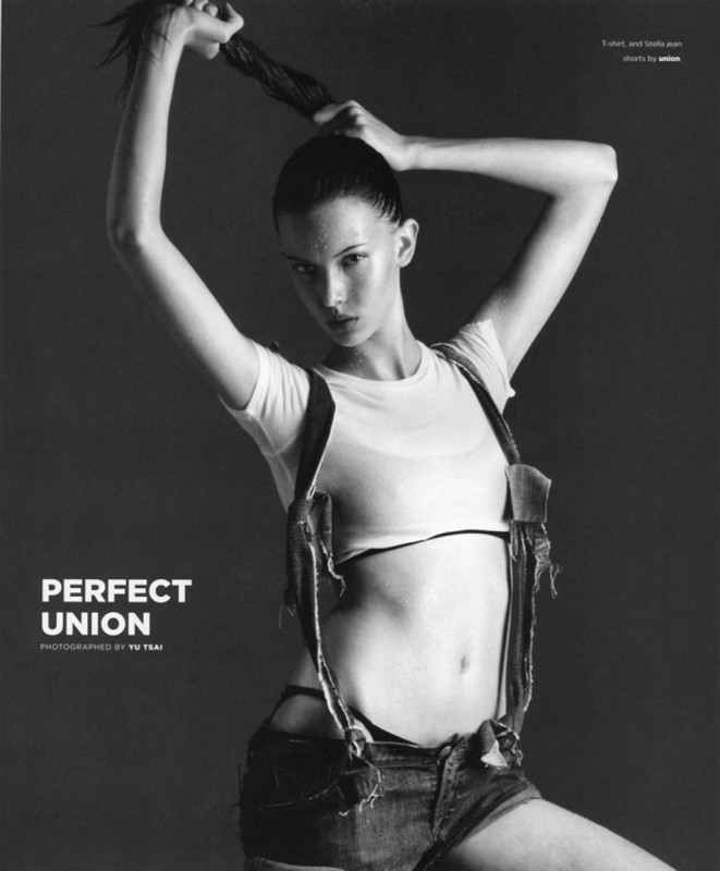 Monochrome portrait of a young woman posing with her hair up, wearing a white top and denim shorts with suspenders against a dark background.