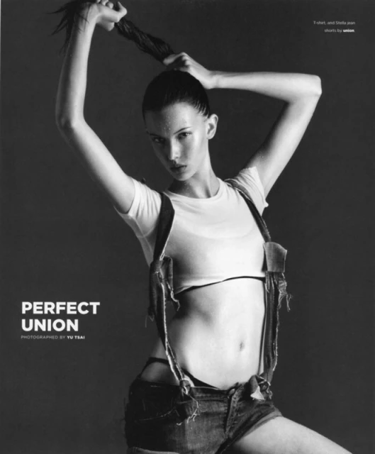 Monochrome portrait of a young woman posing with her hair up, wearing a white top and denim shorts with suspenders against a dark background.