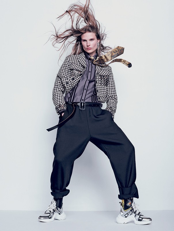 Fashion editorial shot of a female model with long hair, wearing a black and white checkered jacket, black pants and silver chunky sneakers.