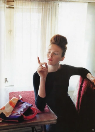 Red-haired woman in a dark top holding up a lipstick tube while seated indoors by a draped window with a patterned bag on the table