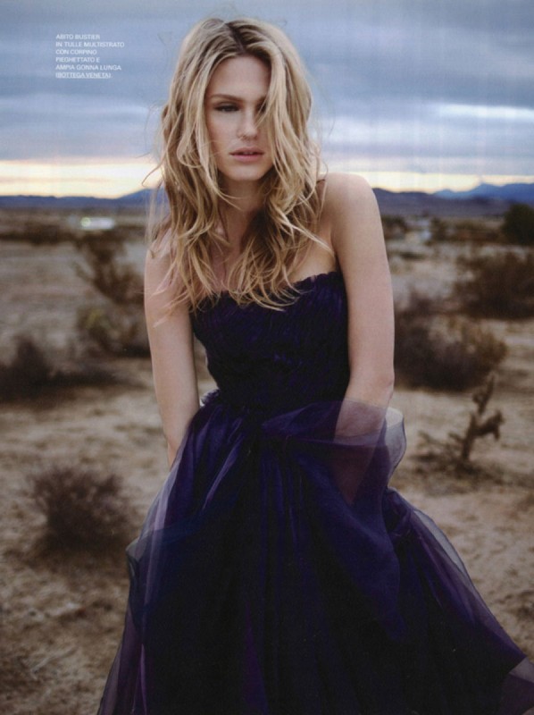 Blonde woman in a strapless dark purple gown standing in an arid desert landscape under an overcast sky.
