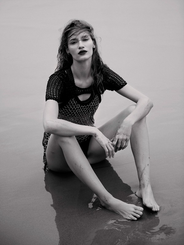 Black and white portrait of a young adult woman with dark lipstick wearing a mesh top while sitting on a sandy beach.