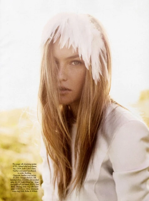 Portrait of a fashionable adult woman wearing a white feather headpiece and long-sleeved top, set against a soft outdoor backdrop.