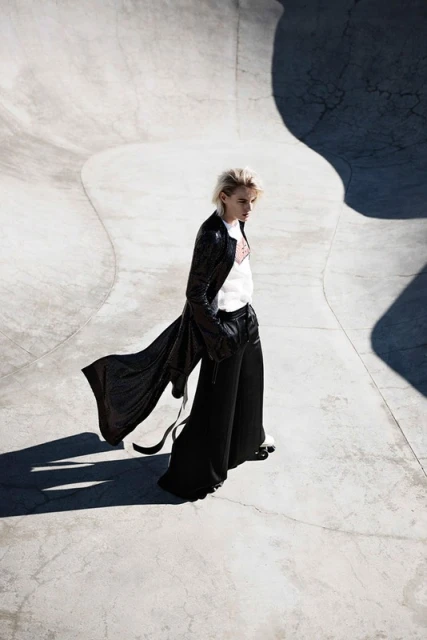 Fashionable woman in long black coat and wide-leg pants walks in a concrete skatepark on a sunny day, captured in a stylish outdoor fashion shot.