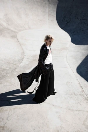 Fashionable woman in long black coat and wide-leg pants walks in a concrete skatepark on a sunny day, captured in a stylish outdoor fashion shot.