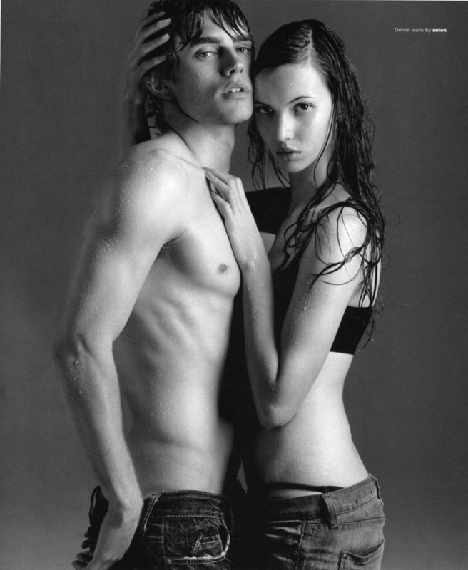 A shirtless man and a woman in jeans posing together with wet-look hair against a neutral grey studio background.