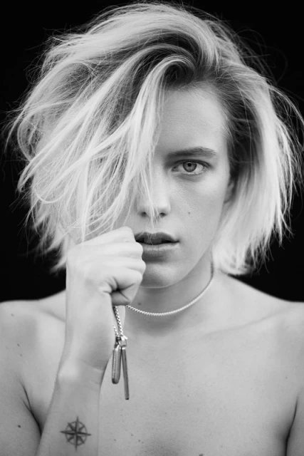 Black and white portrait of a person with tousled blond hair, holding a dog tag necklace, set against a stark black backdrop.